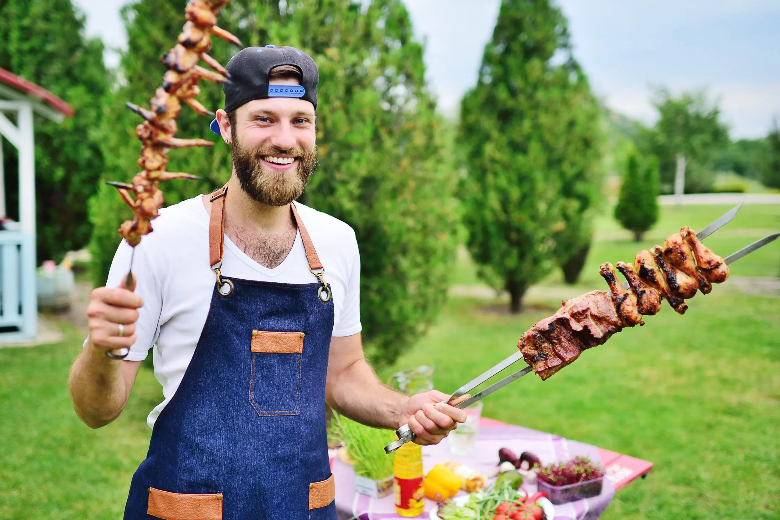 young bearded man in a cap and denim apron smiles and holds a skewer with delicious barbecue meat cooked on the grill against the background of greenery, a Park and a gazebo. Butchery with Catering